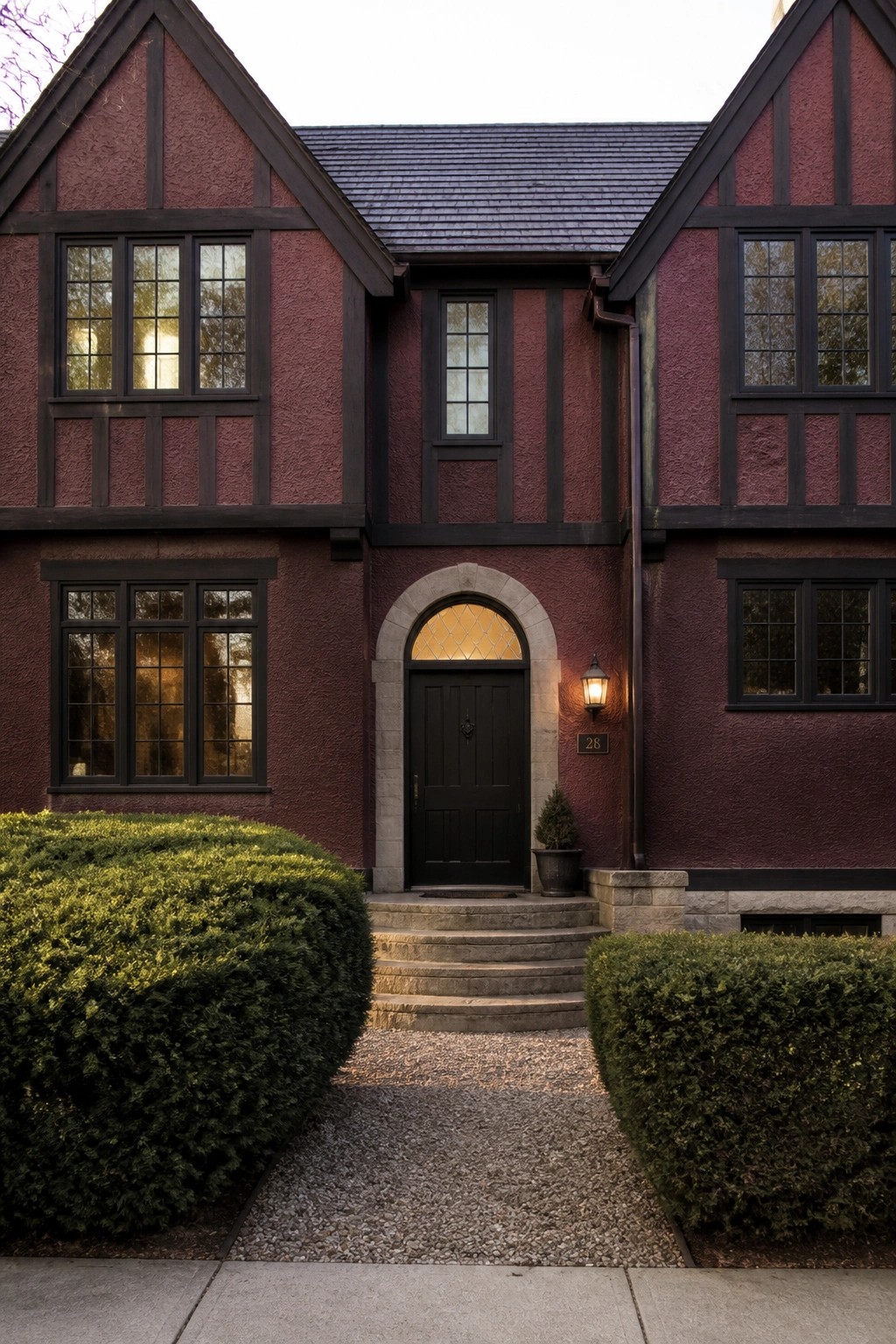 Red Tudor house with black door, stone arch, steps, and green shrubs.
