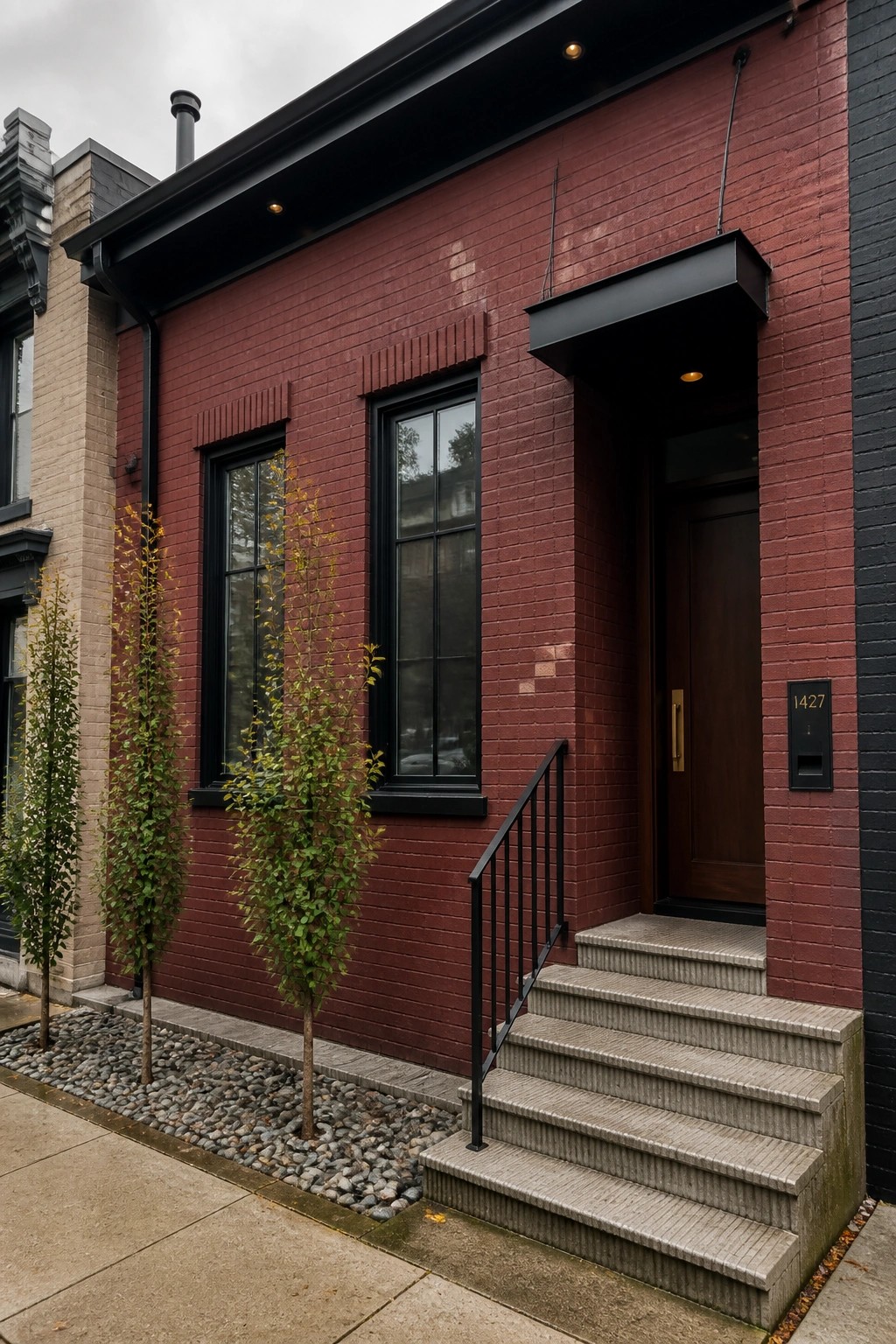 A maroon brick house with black window frames, a dark wood door, concrete steps, and two small trees in front.