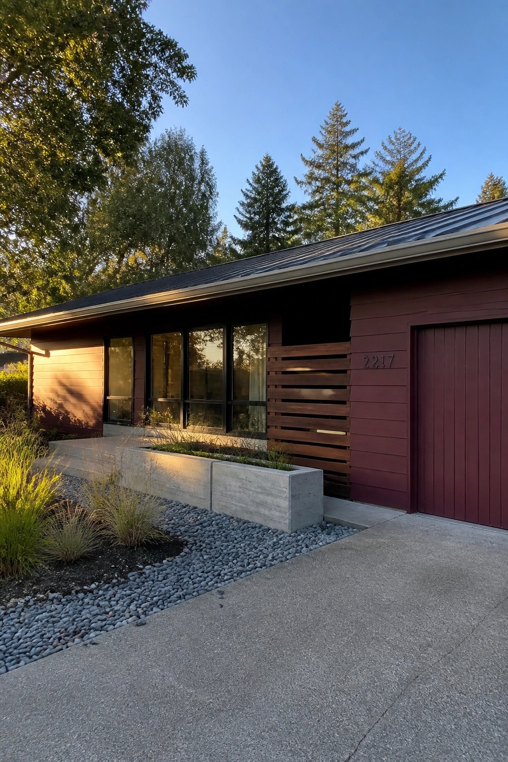 A single-story modern house with deep maroon horizontal siding, black-framed windows, a matching garage door, and concrete planters along the front.