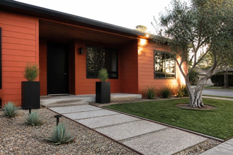 Orange wood-sided house with matte black door and window frames, concrete steps, and gravel landscaping with agave plants.
