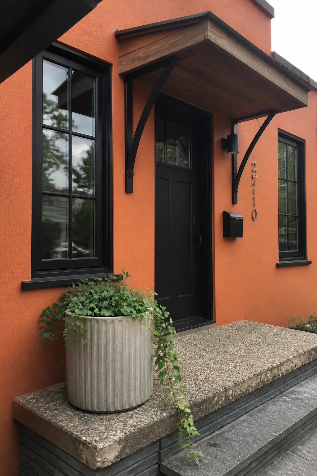 A front view of an orange stucco house with matte black window frames, a black door, a wooden entry overhang with black brackets, and a concrete planter on the steps.