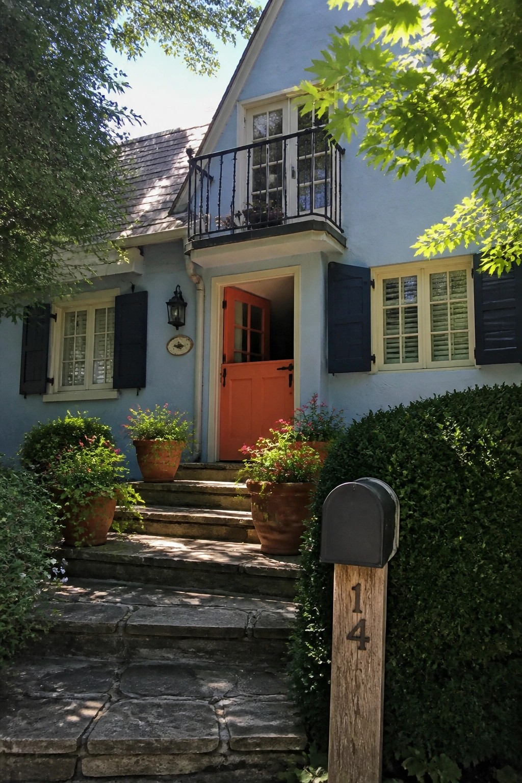 Blue house with orange door, stone steps, potted plants, and mailbox marked 14