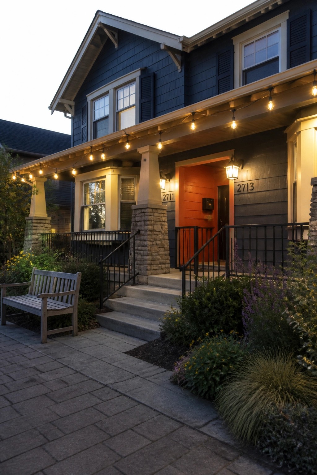 Dark blue house with orange door, string lights on porch, wooden bench and garden.