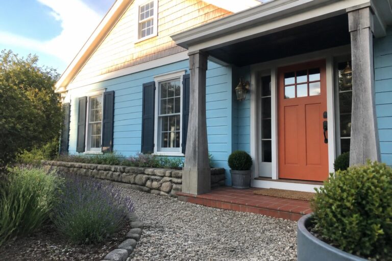 A light blue house exterior with navy shutters, an orange front door on a covered porch with stone columns, and gravel landscaping with lavender and shrubs in the foreground.