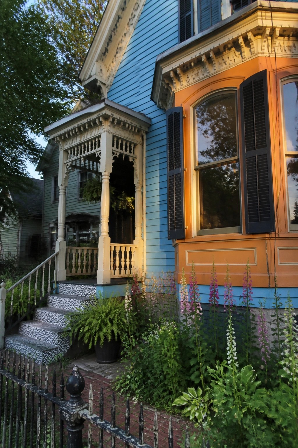 A two-story house with light blue siding, orange painted trim on a bay window, navy shutters, and a white front porch with steps leading up from a brick path.
