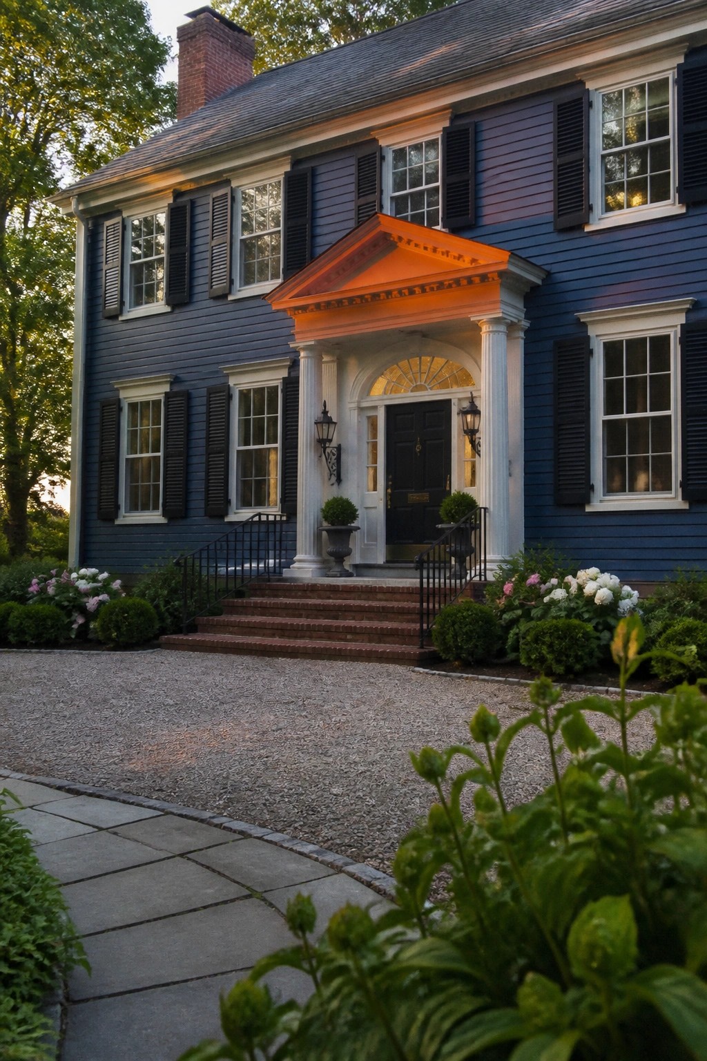 Blue two-story house with glowing orange portico, black shutters, brick steps, and gravel drive.