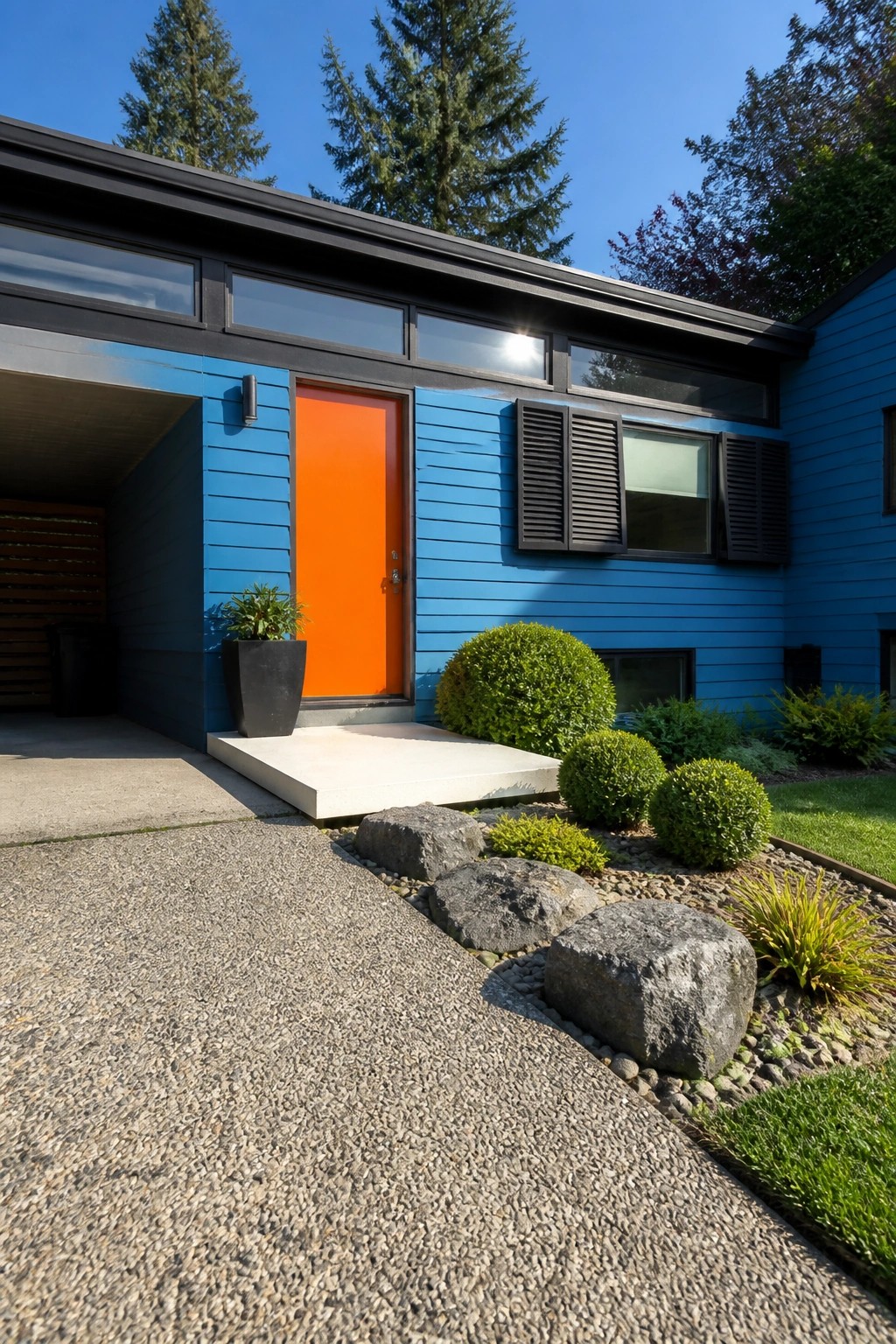 A blue house exterior with an orange front door, navy shutters, a concrete step, and rocks along the driveway.