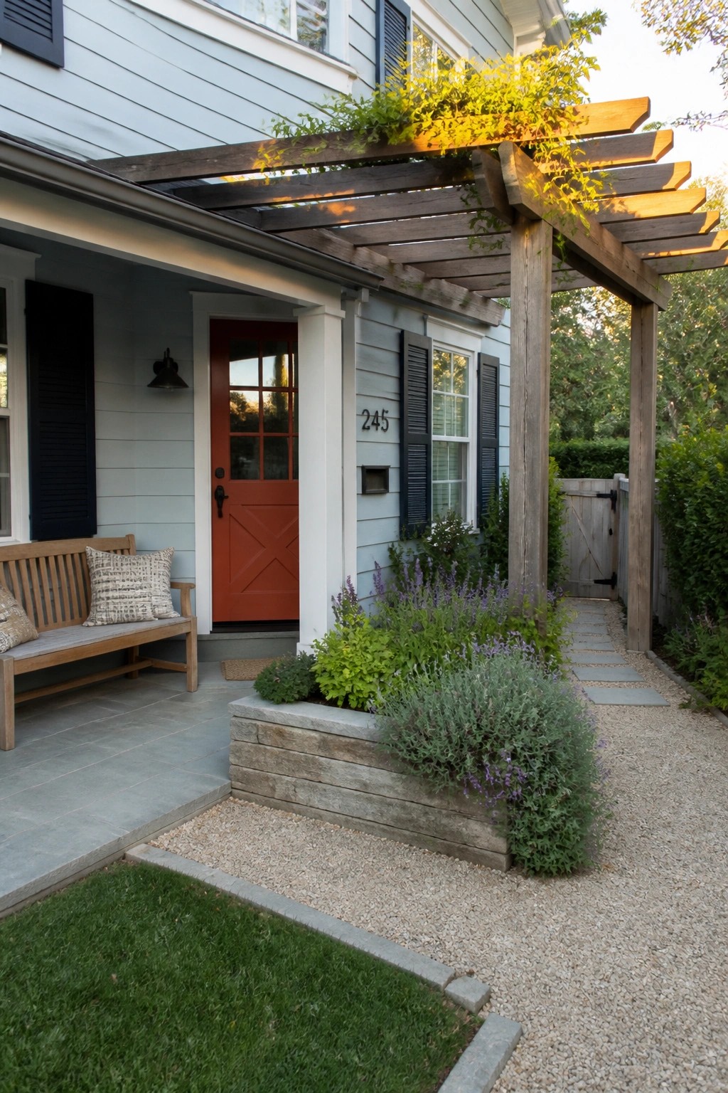 Light blue house exterior with an orange front door, navy shutters, wooden pergola, and gravel path with plantings.