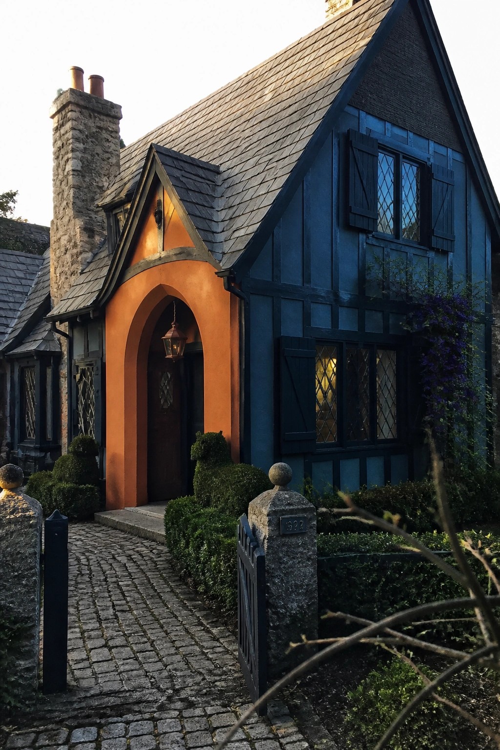 A blue house with navy shutters and an orange arched entryway beside a cobblestone path and stone chimney.