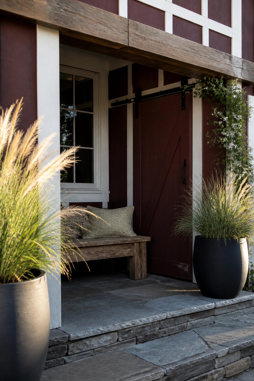 Exterior view of a maroon farmhouse entry featuring white board-and-batten trim, a sliding barn door, a wooden bench with pillows, and large potted grasses on stone steps.
