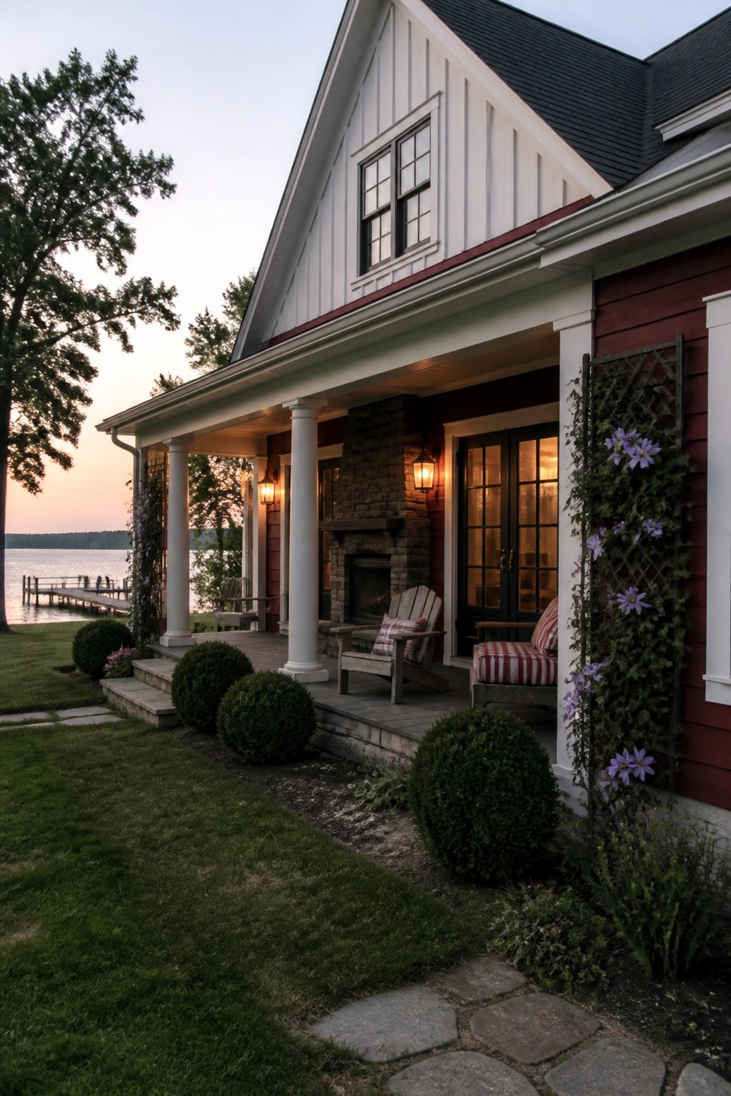 A two-story maroon farmhouse with white board-and-batten siding on the upper gable, a stone chimney, white porch columns, and a covered front porch facing a lake.