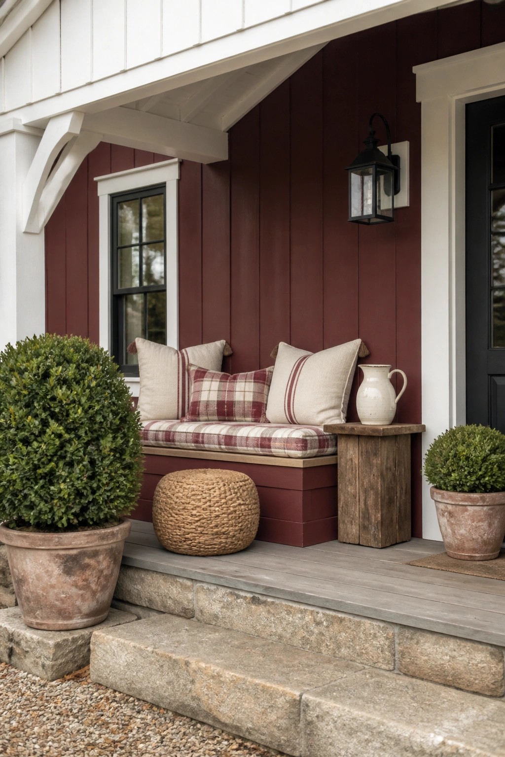 A maroon farmhouse exterior with white trim shows a built-in porch bench with cushions, a black lantern on the wall, and potted shrubs on stone steps.