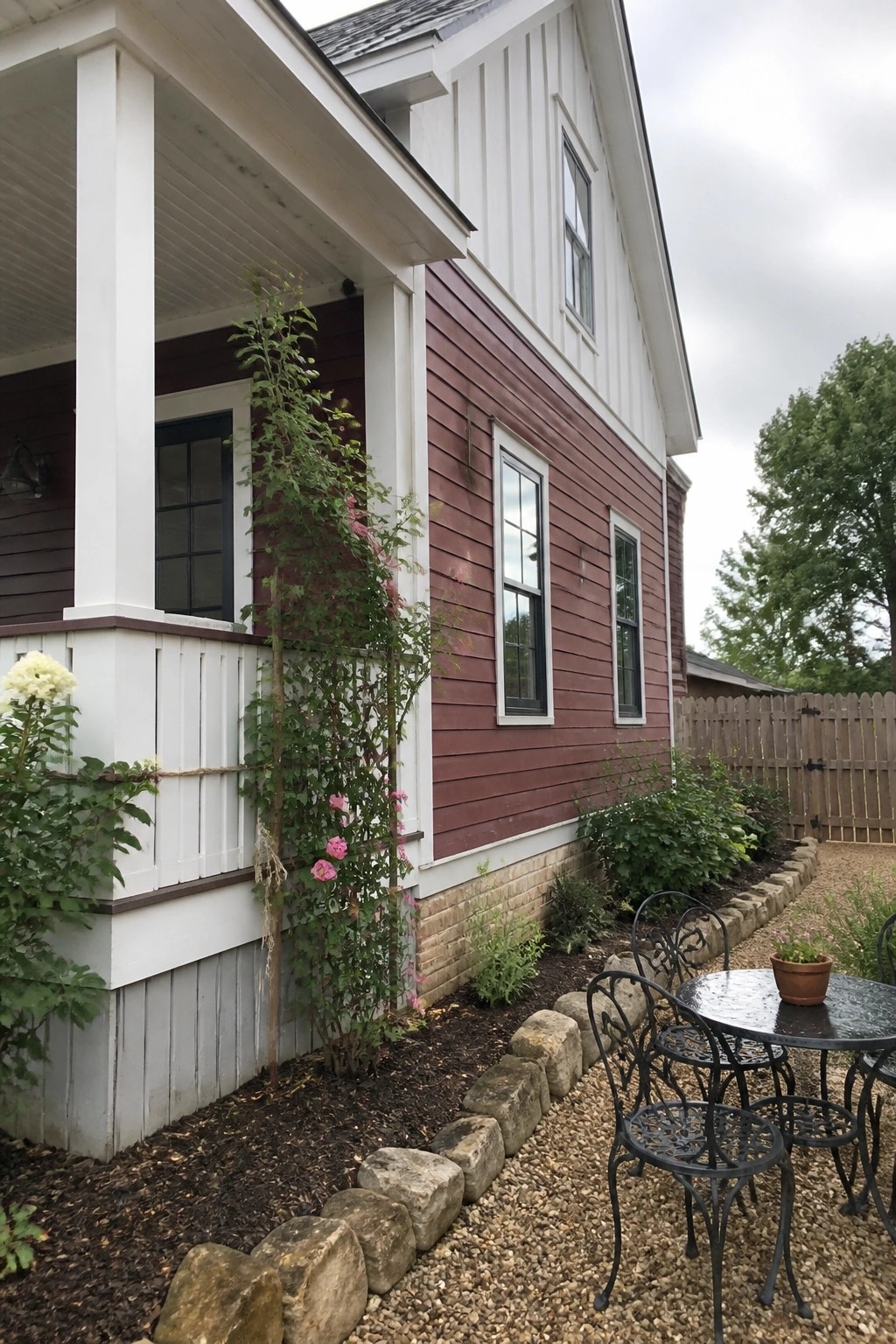 A two-story maroon farmhouse with white board-and-batten on the porch and gable ends, black window trim, a gravel side yard with black metal patio furniture, and a rose trellis along the foundation.