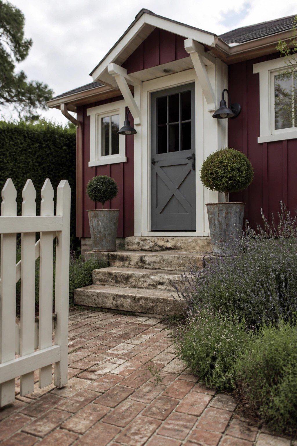 A maroon house exterior with white trim around windows and door, a gray front door, stone steps, and potted shrubs beside a brick walkway.