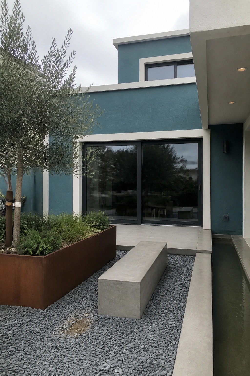 A modern house with teal exterior walls, white trim, large black-framed sliding doors, and a gravel courtyard with a concrete bench and metal planter.