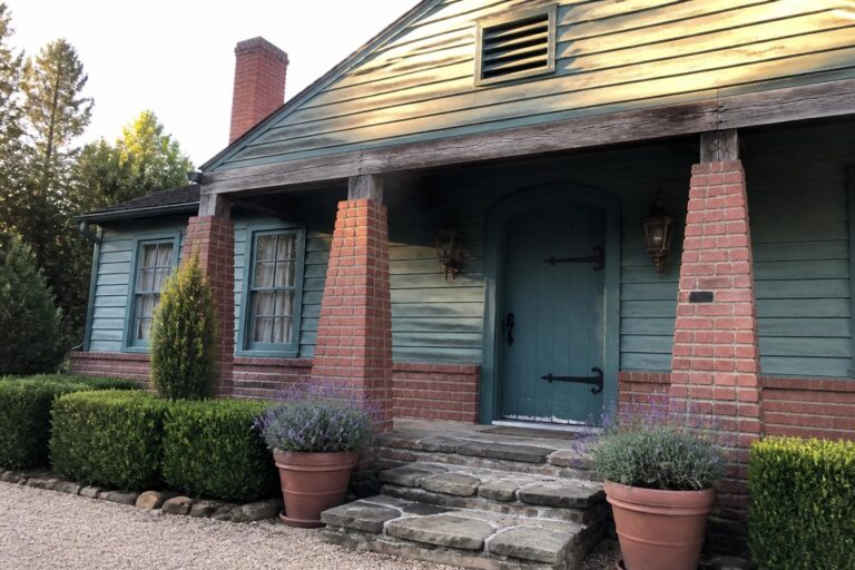 A small teal house with red brick columns framing the front porch, a teal arched door, stone steps, and potted lavender plants.