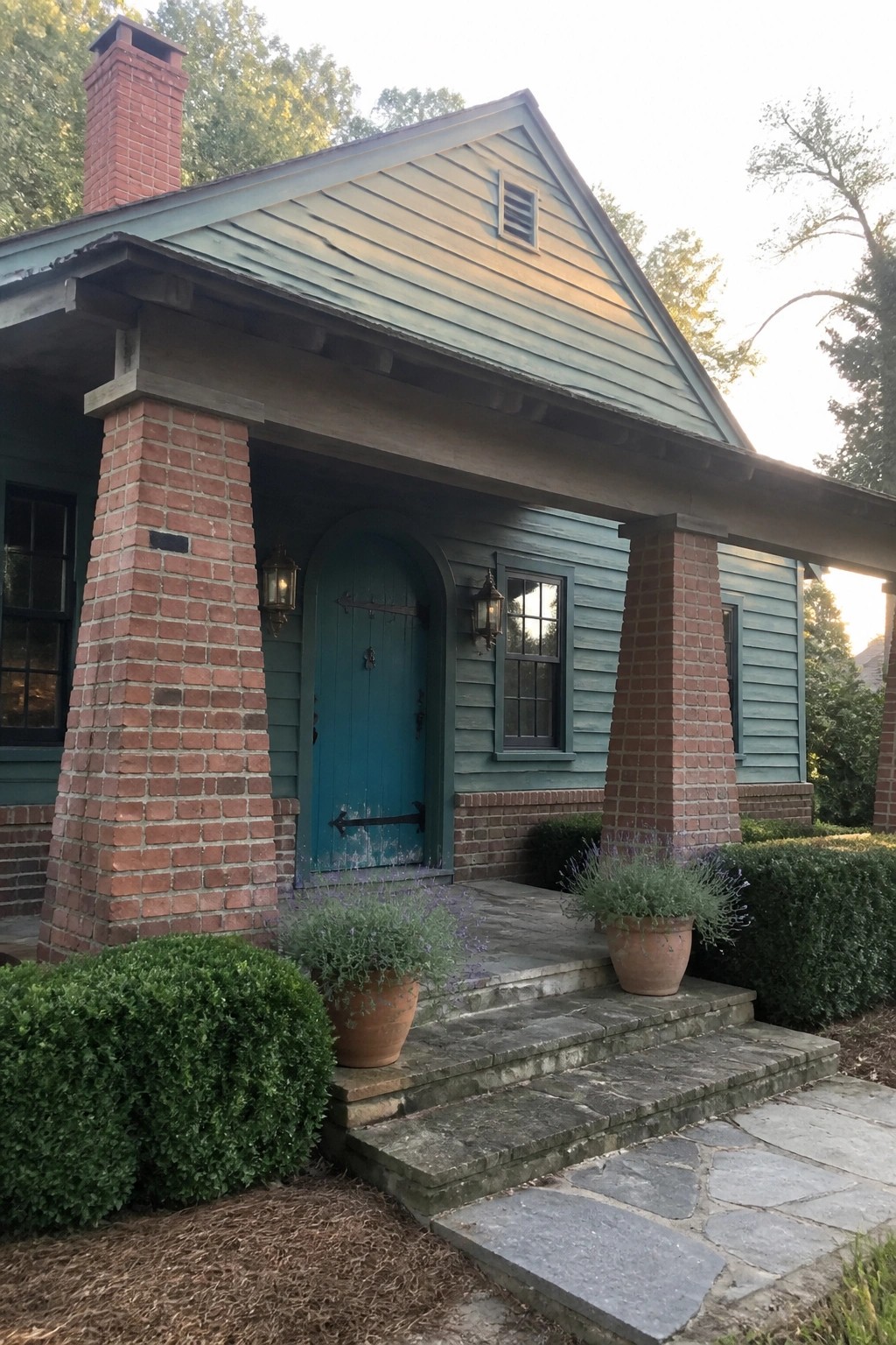 A small teal house with red brick columns framing the front porch, a teal arched door, stone steps, and potted lavender plants.