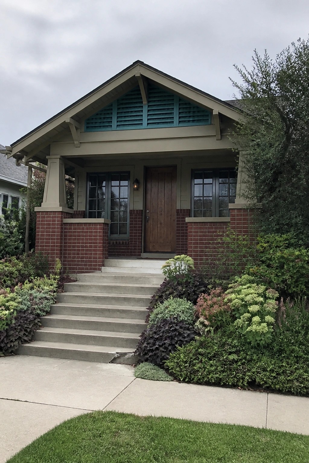 Beige bungalow with teal vented gable, wooden door, brick pillars, and concrete steps.