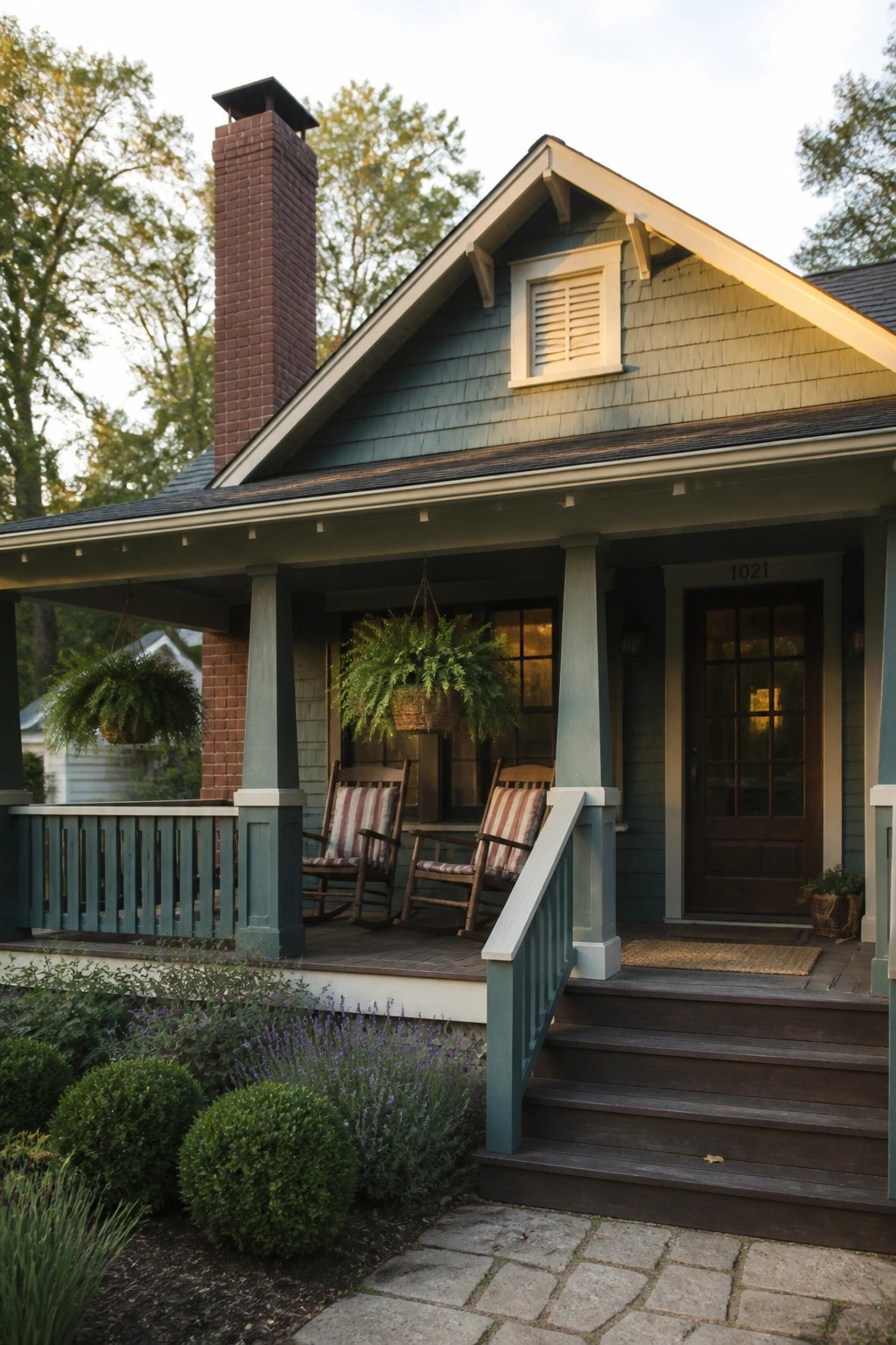 A teal house with red brick chimney, covered porch holding two rocking chairs, and steps leading up to the front door.