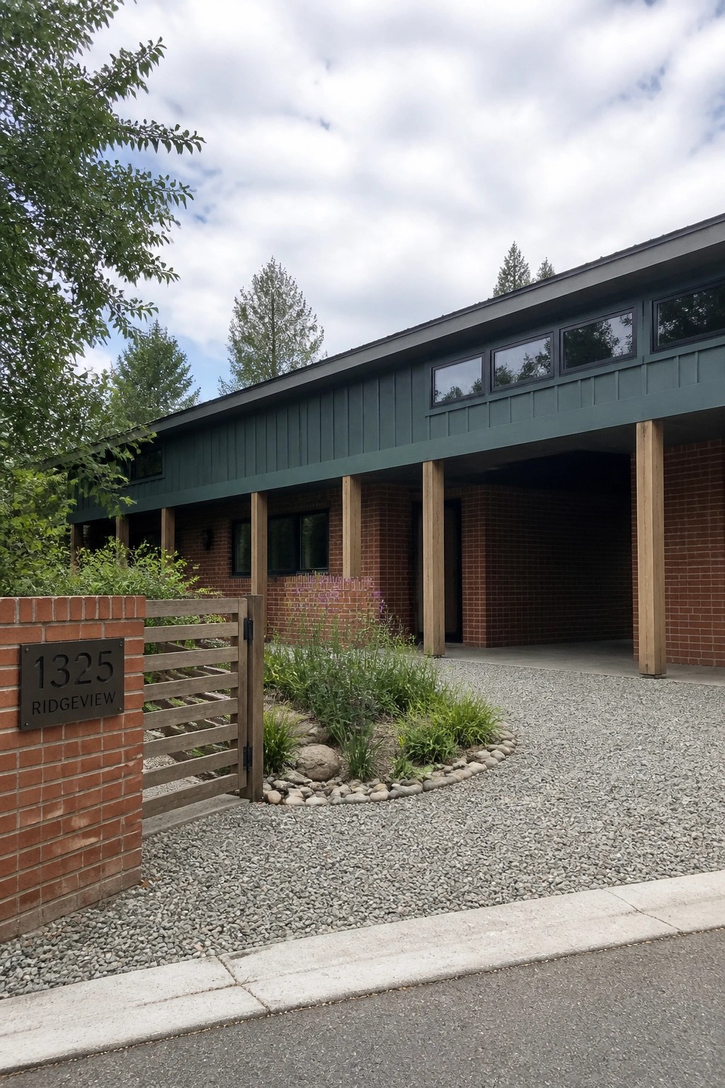 A red brick house with teal siding above, wooden support posts under a covered carport, a gravel driveway, and a brick pillar with an address sign at the front.