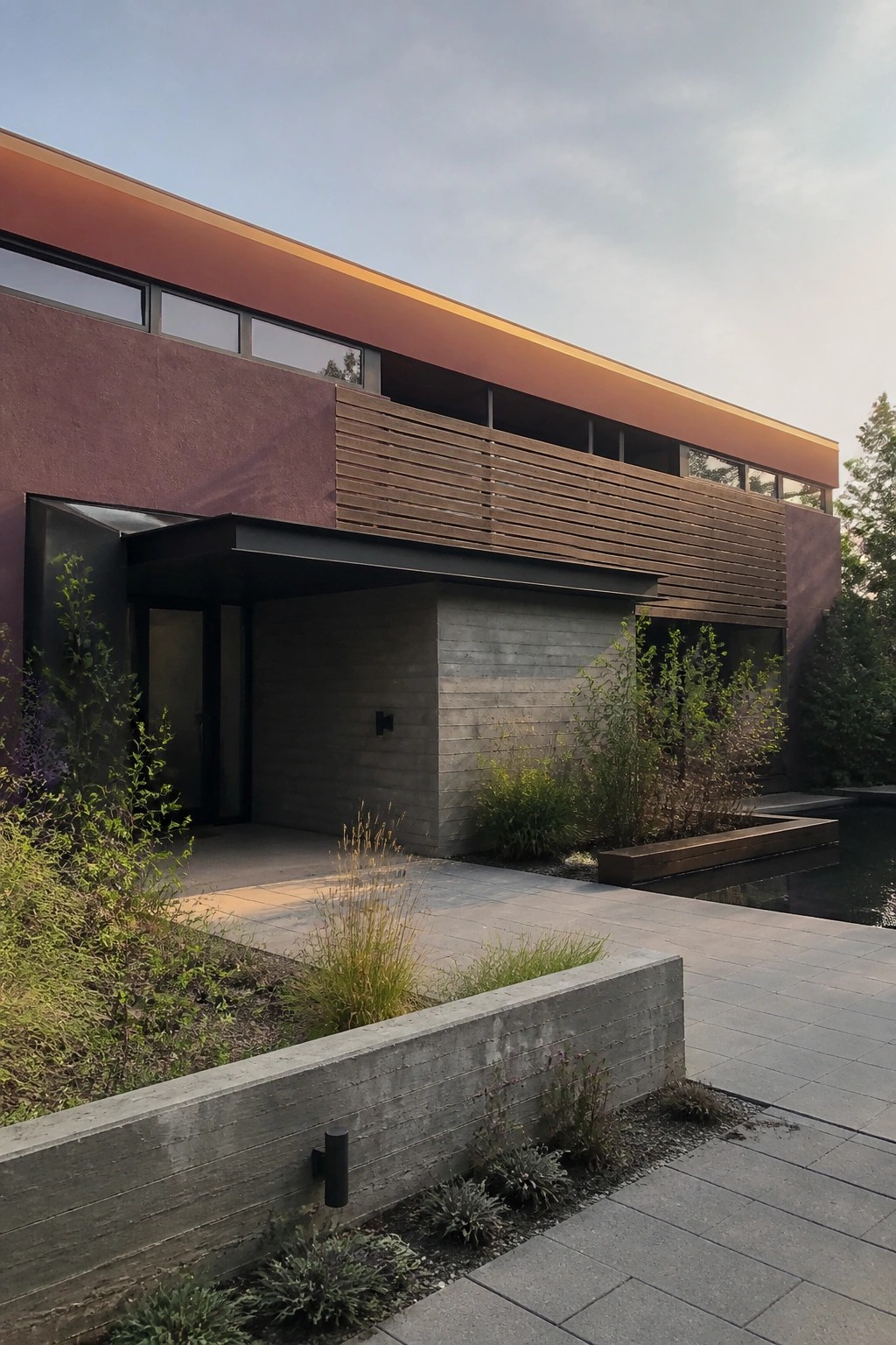 Modern two-story house with flat roof, burgundy panels, horizontal wood louvers, large windows, and wood front door above concrete steps.