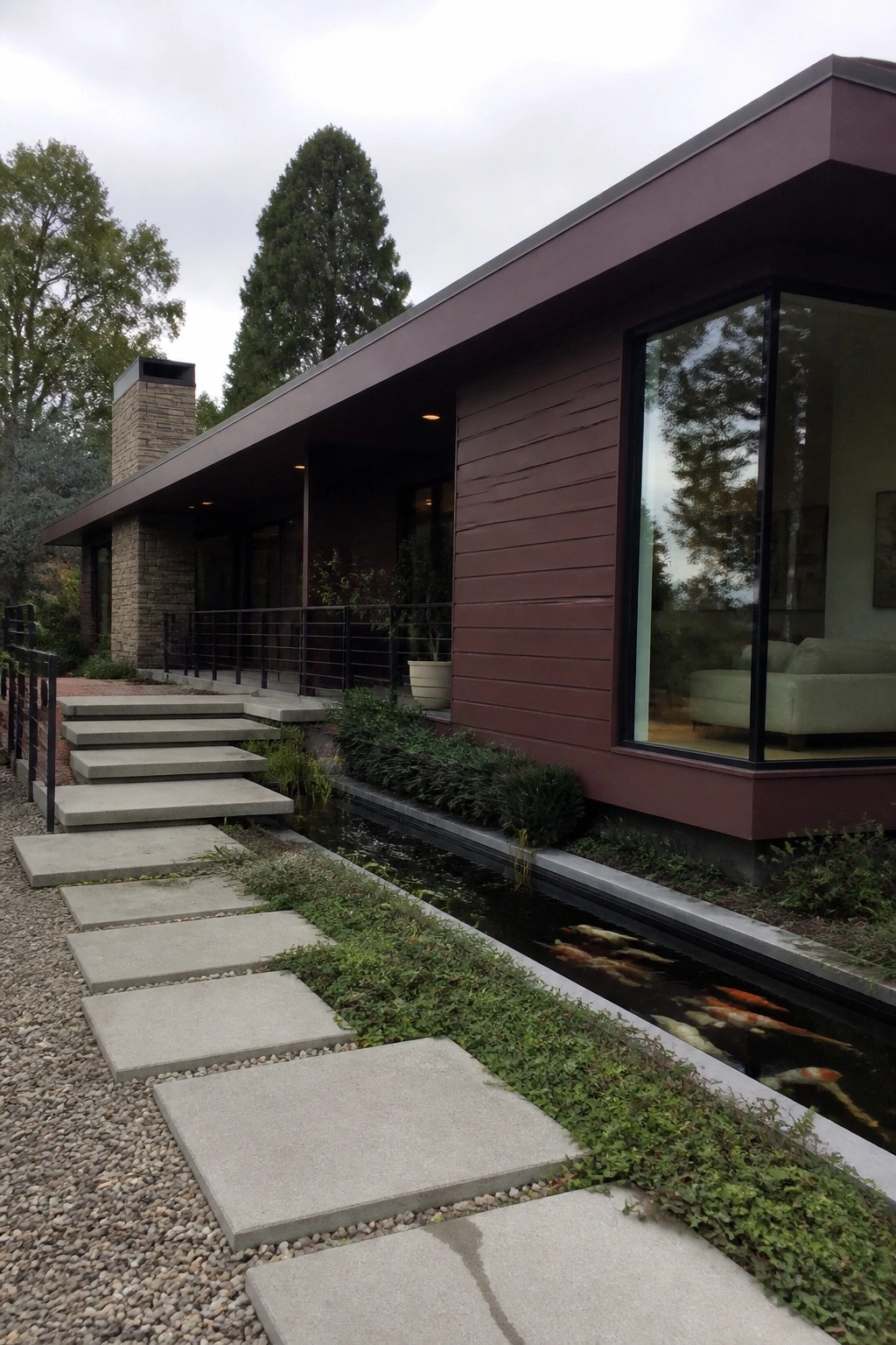 A modern single-story burgundy house with a flat roof, stone chimney on the left, concrete stepping stones leading to the entrance, and a narrow water feature with fish running along the right side.