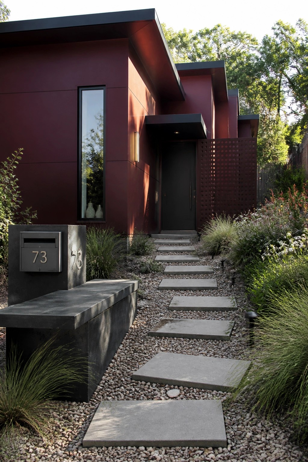 A modern burgundy house with stepped flat rooflines has a gravel path with concrete pavers leading to a black front door, with a metal mailbox labeled 25 in the foreground.