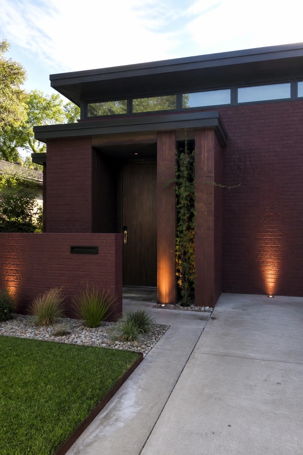 Modern dark red brick house with wooden door, ivy and concrete walkway.