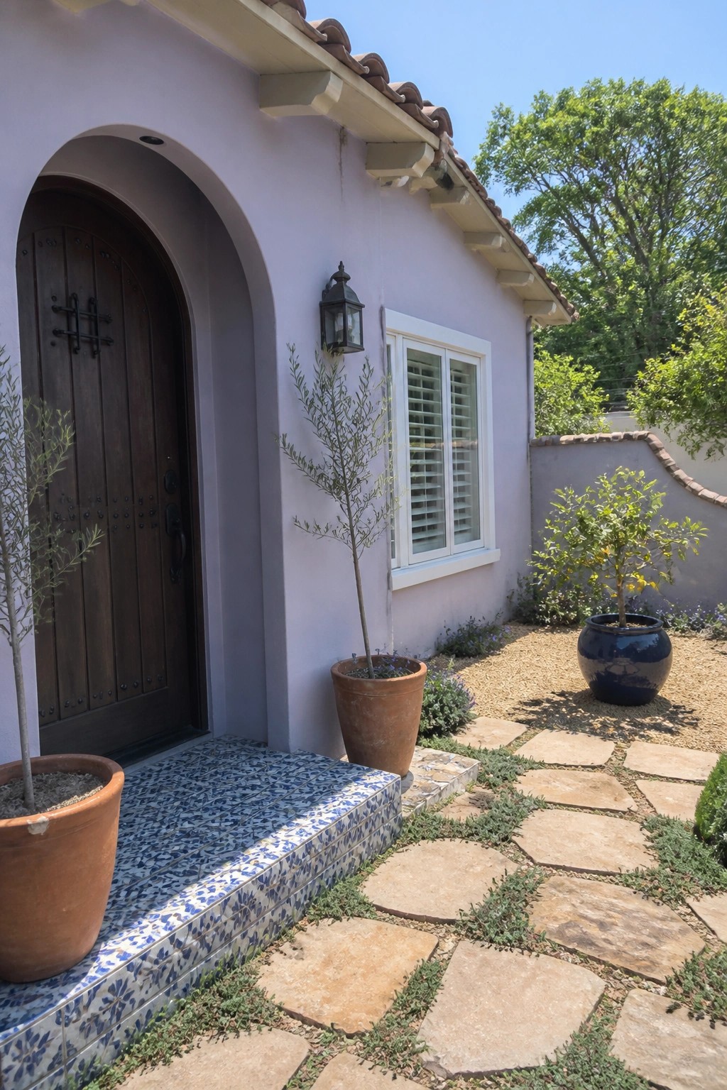 A light purple stucco house exterior with a dark wooden arched door, a window fitted with white shutters, and potted plants beside a stone pathway.