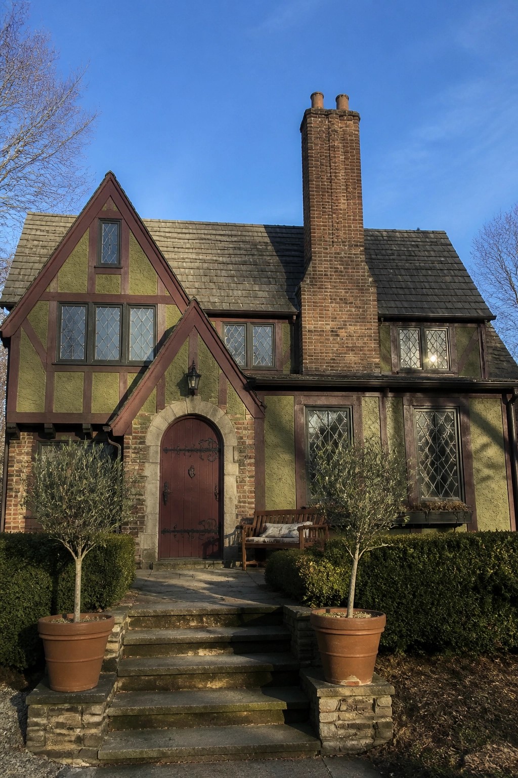 A two-story Tudor-style house with green stucco walls, maroon half-timbering, a red front door, brick chimney, and potted olive trees beside the front steps.