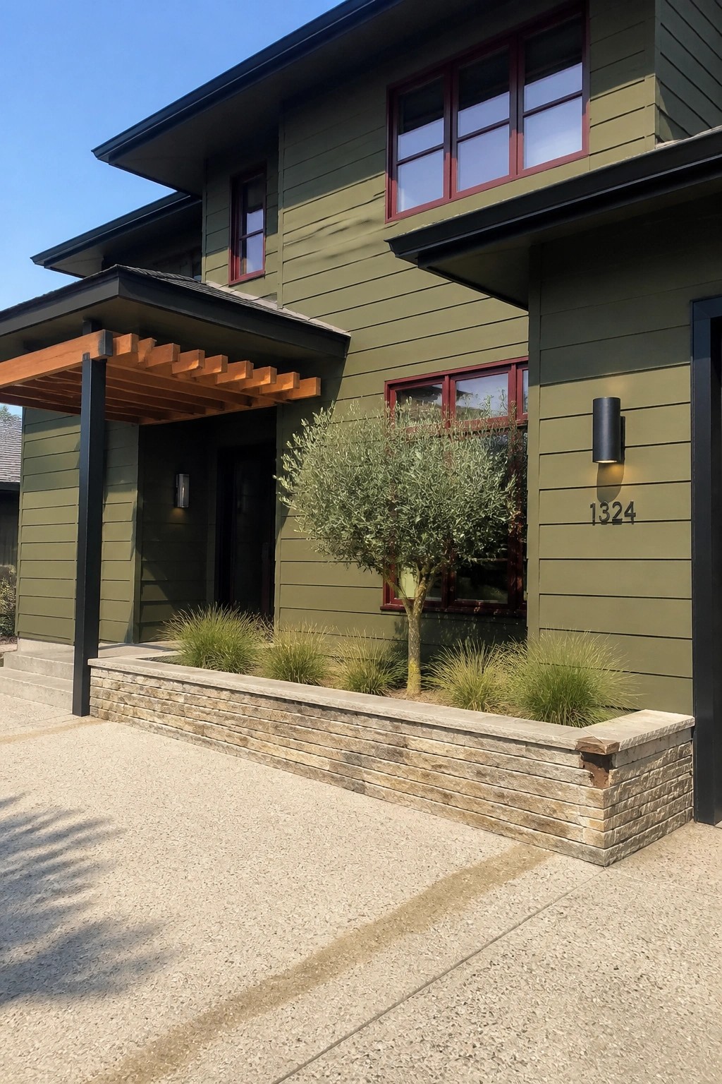 A two-story house with olive green siding, maroon window frames, a stone planter bed holding an olive tree and grasses, and a wooden pergola over the front entrance.