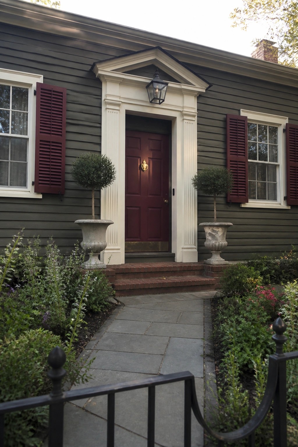 Front view of a house with olive green siding, a central maroon door framed by white columns and a pediment, maroon shutters on flanking windows, and a stone path leading to brick steps.