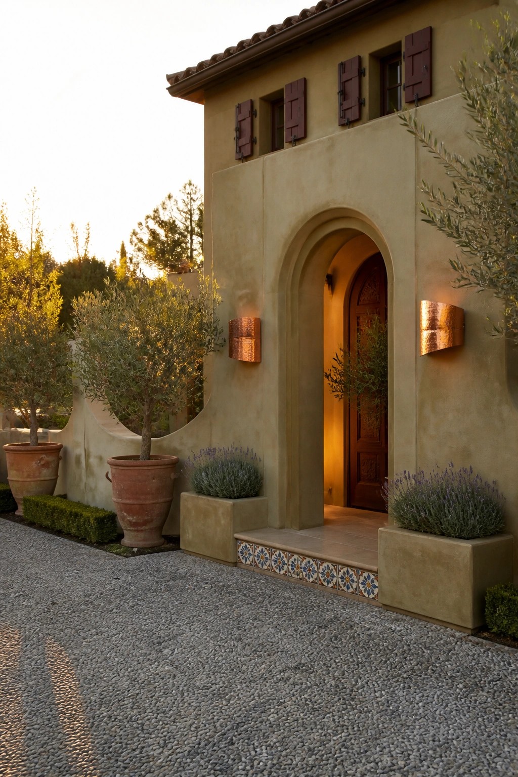 A stucco house with an arched entry, maroon shutters, copper sconces, and potted olive trees next to a gravel driveway.
