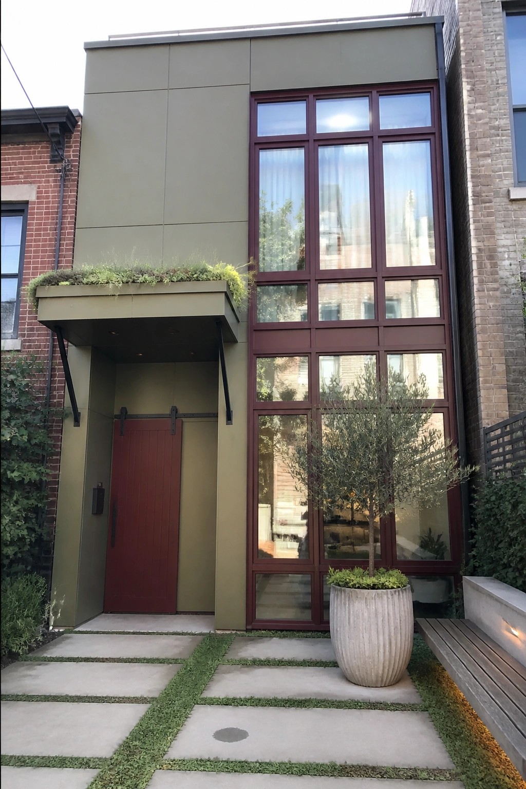 Modern house exterior with olive green panels, maroon window frames, and a red door beneath a green canopy.