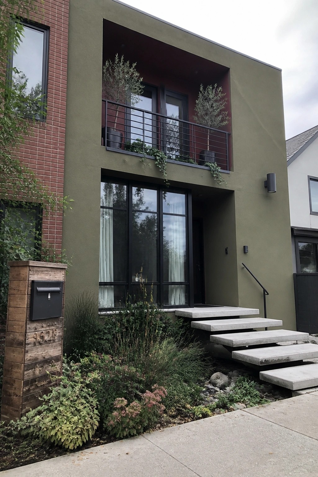 A two-story modern house with olive green stucco walls, a recessed maroon balcony with black metal railing, black-framed windows, and concrete stairs leading to the entry.
