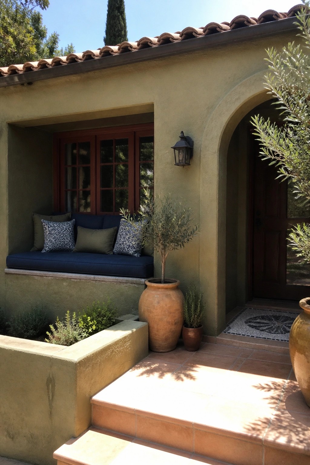 Green stucco house with arched doorway, blue-cushioned window seat, and terracotta planters.
