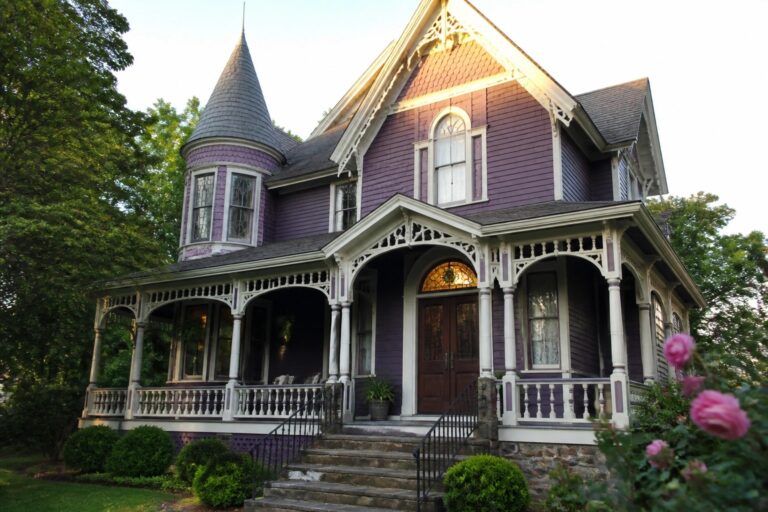 18 Classic Purple Victorian House Exteriors Featuring Ornate White Trim Purple Victorian house with turret, ornate white porch, stone steps, and pink roses.