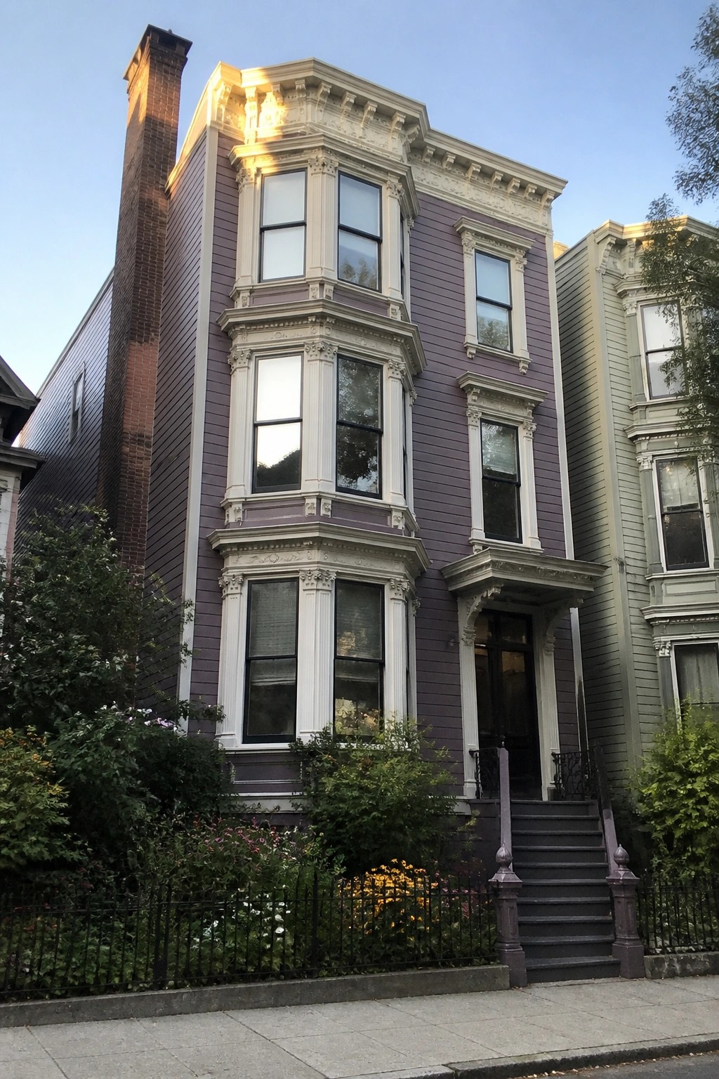 A purple Victorian house with white trim on the bay windows and entrance, shown from the sidewalk with dark steps leading to the front door.