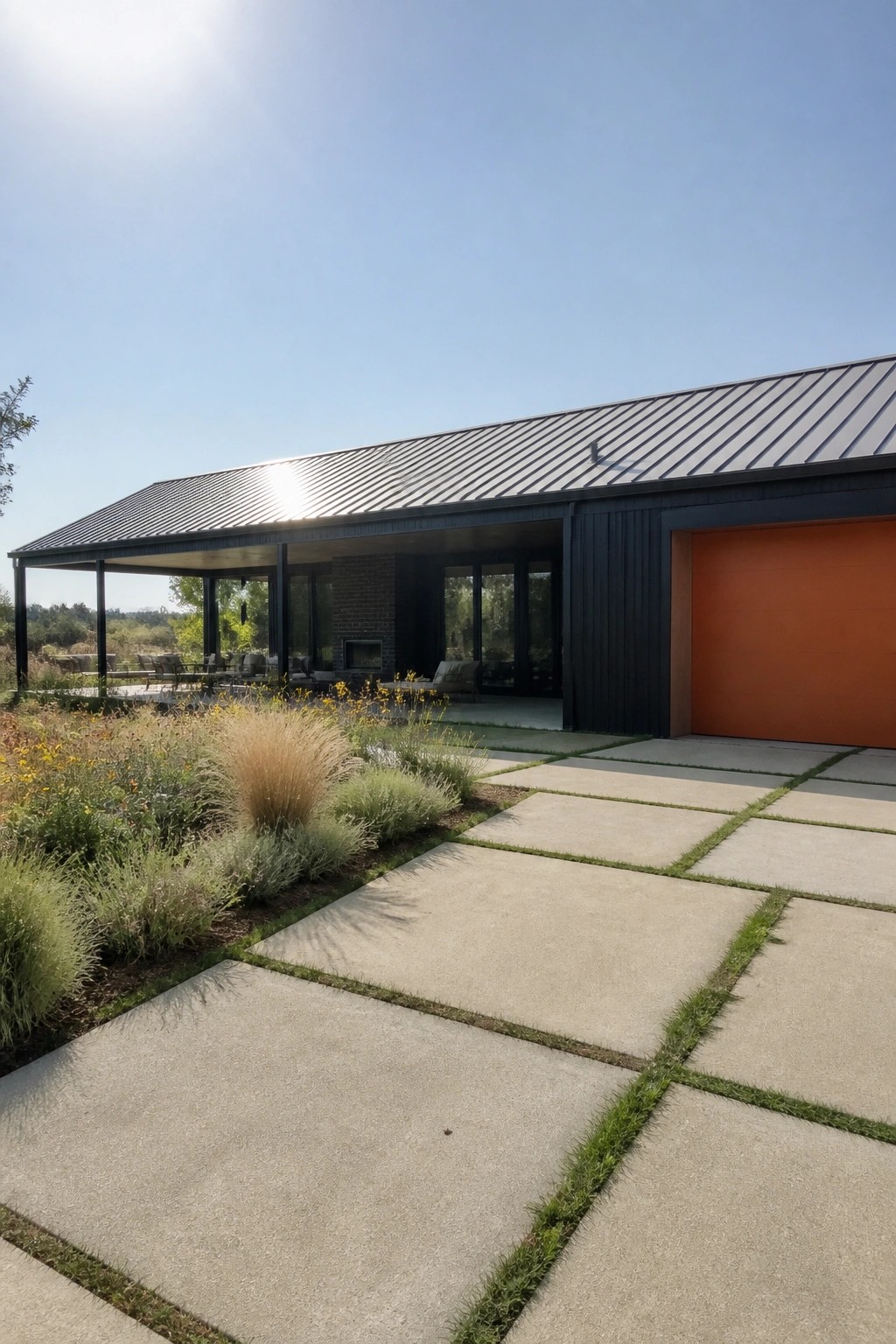 A modern single-story house with black siding, charcoal metal roof, bright orange garage door, and open covered patio next to concrete pavers and landscaping.