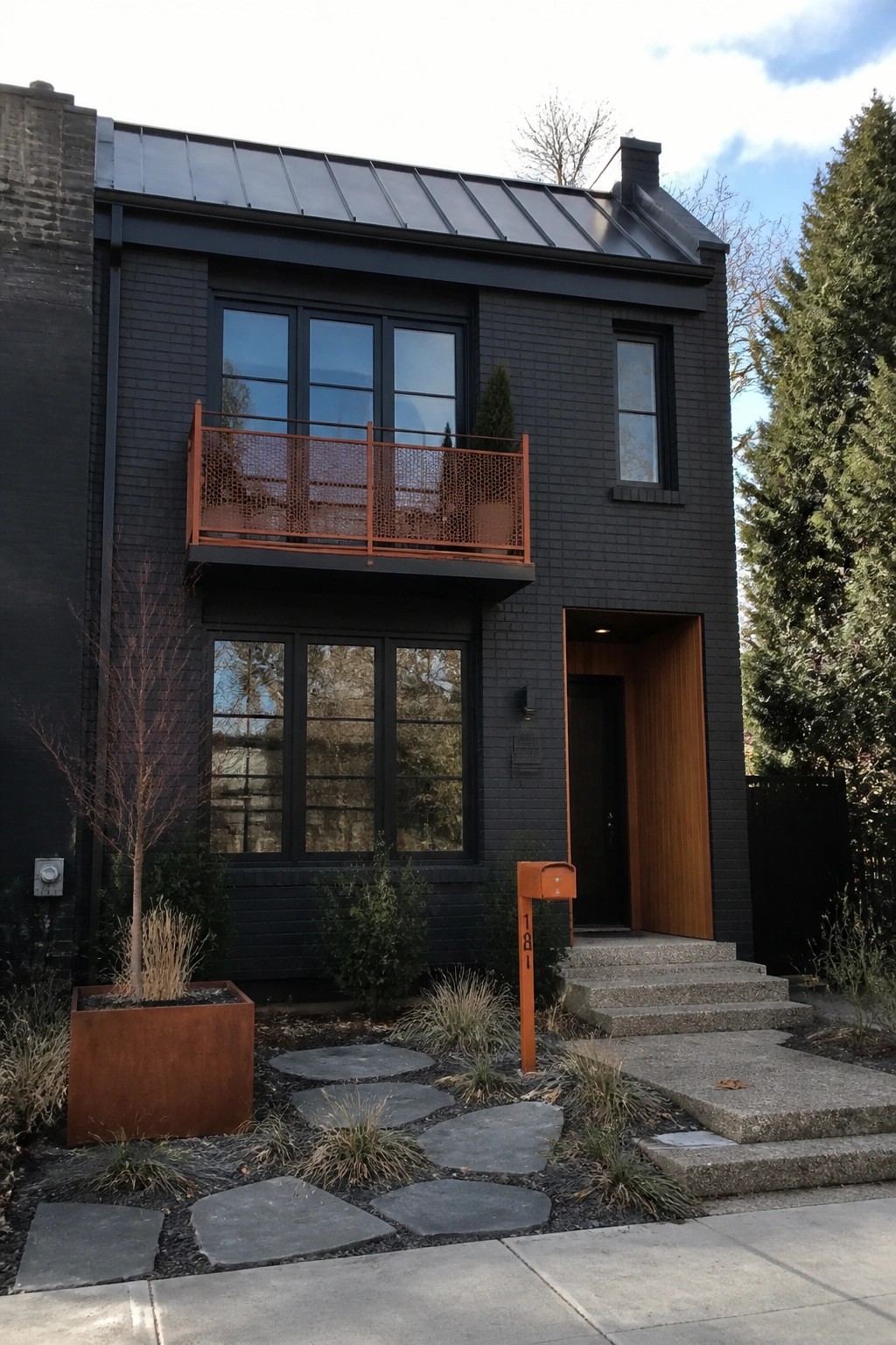 A two-story black brick house with a charcoal metal roof, an orange mesh balcony railing on the second floor, an orange mailbox, and a matching orange planter in the front yard.