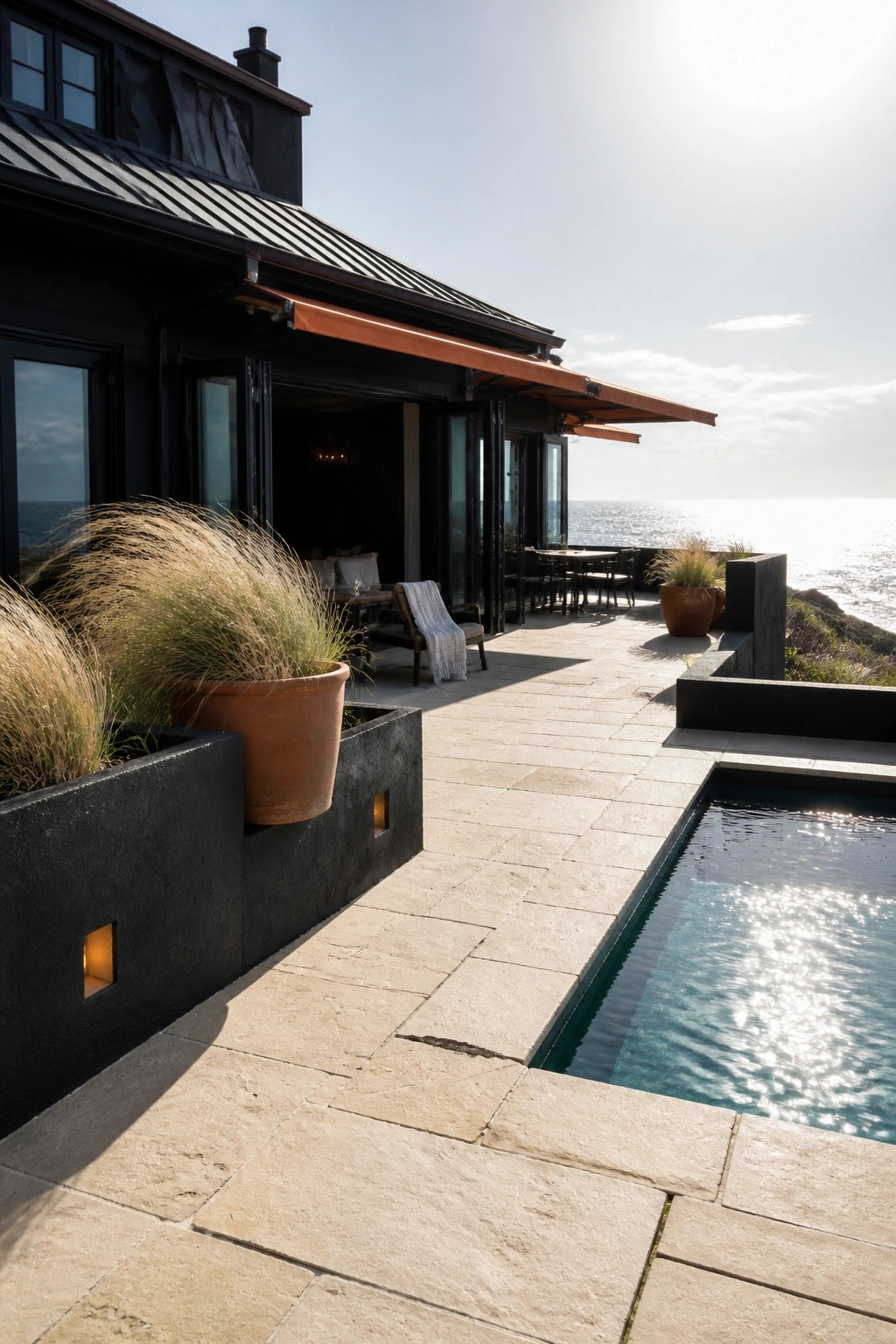 A modern black house with charcoal metal roofing has a large stone patio with an integrated pool, orange retractable awnings, terracotta planters, and seating areas facing the ocean.