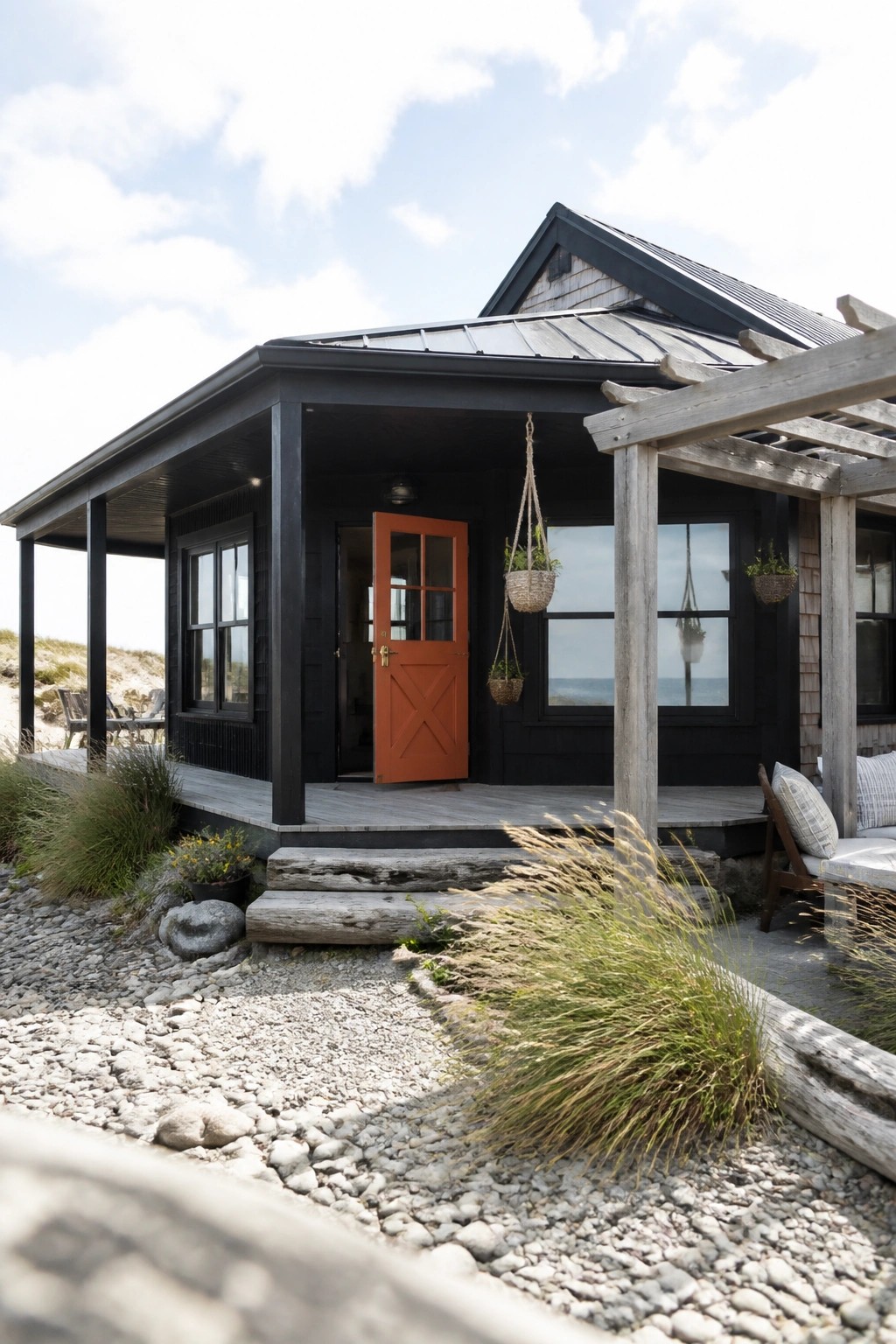 A black house with an orange front door, charcoal metal roof, and wooden porch set on a gravel area near the beach.