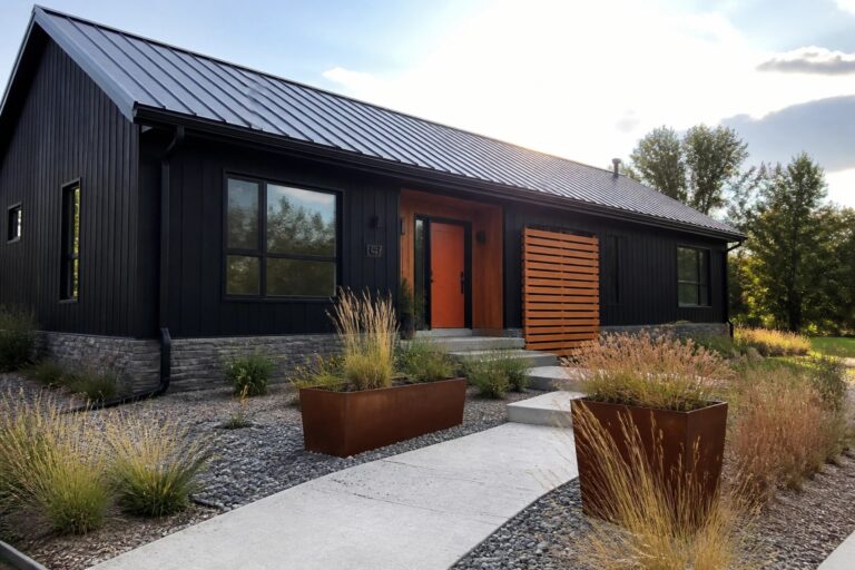 A front view of a modern house with black vertical siding, an orange door, a charcoal metal roof, and rusted metal planters along a concrete walkway.