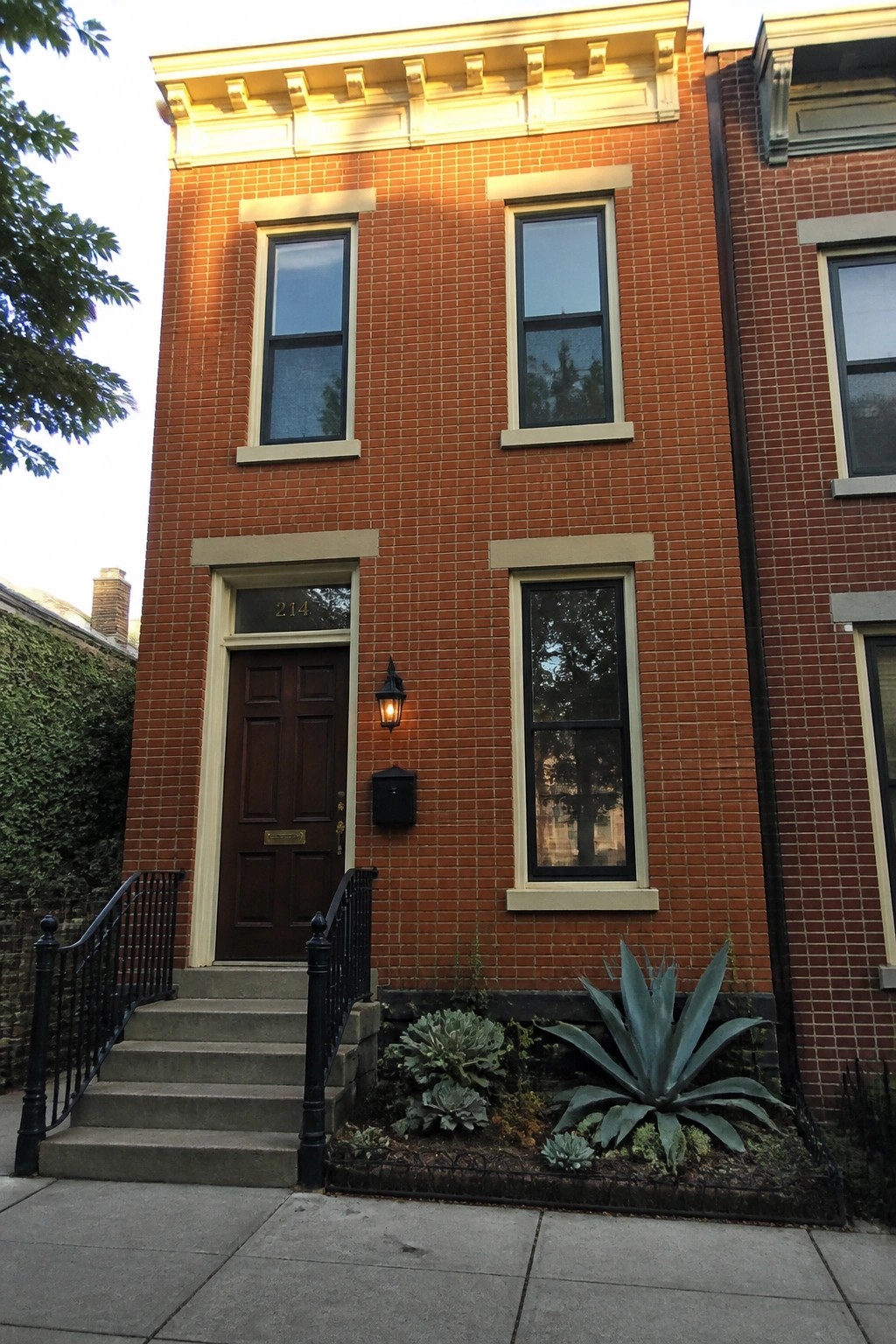 A narrow two-story brick row house with burnt orange bricks, cream trim around the windows and door, a dark wood front door, and a small front garden bed.