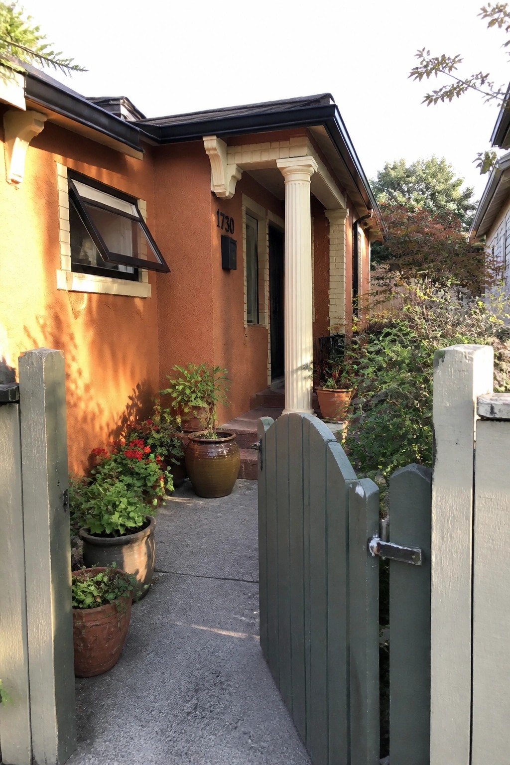 A burnt orange stucco house with cream trim and columns, seen through an open wooden gate with potted plants along the path.