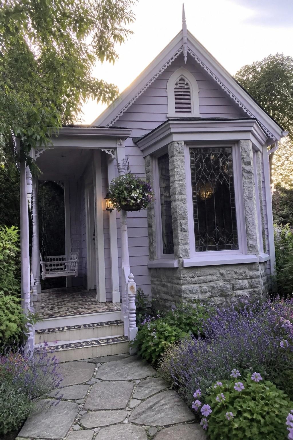 A light lavender house exterior featuring gray stone on the lower walls and bay window, a white porch with a hanging swing, and lavender plants along a stone path.