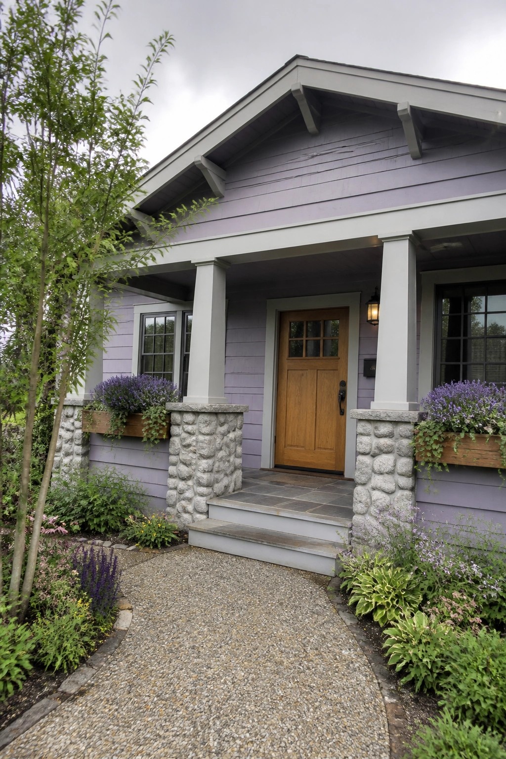 A house with light purple siding, a wooden front door, stone porch pillars, and window boxes with purple flowers beside a gravel path.