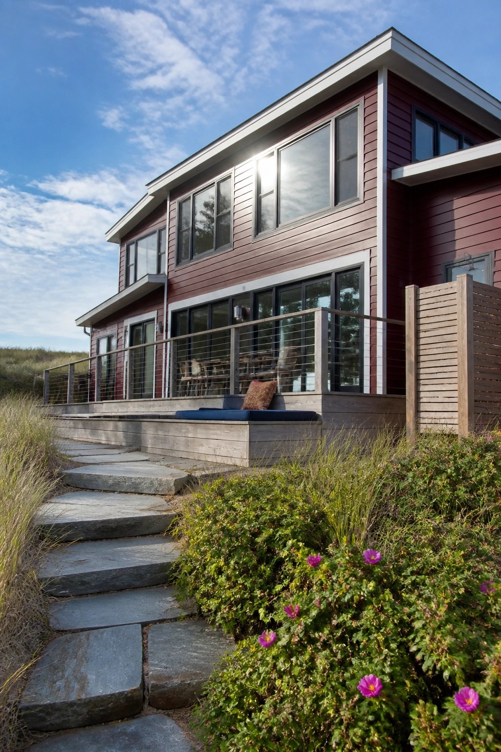 Modern maroon house with large windows, deck, stone steps, and purple flowers.
