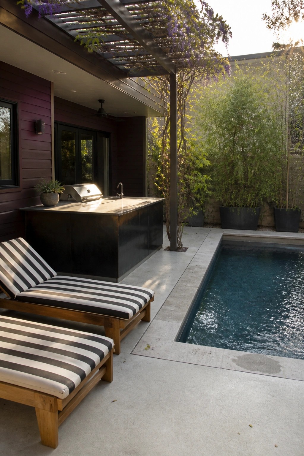 A burgundy house exterior with smooth siding next to an outdoor kitchen, two striped lounge chairs, and a rectangular pool on a concrete patio.