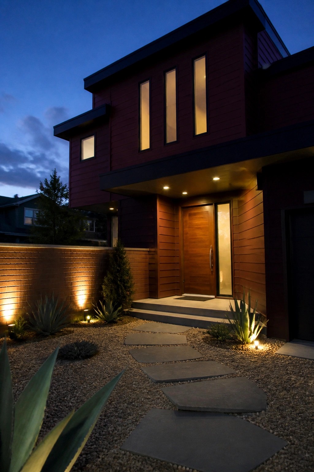 Modern red wooden house with glowing windows, wooden door, and stone path at dusk